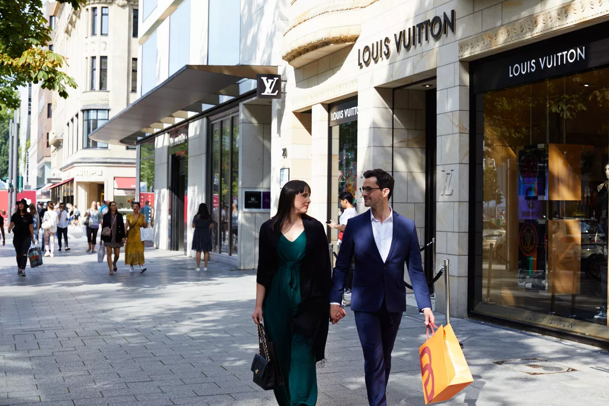 Ein Paar das auf der Könisgallee in Düsseldorf einkaufen geht. Im hintergrund die Fassade von Louis Vuitton sowie der Königsallee mit einigen Menschen die ebenfalls dort entlang spazieren.
